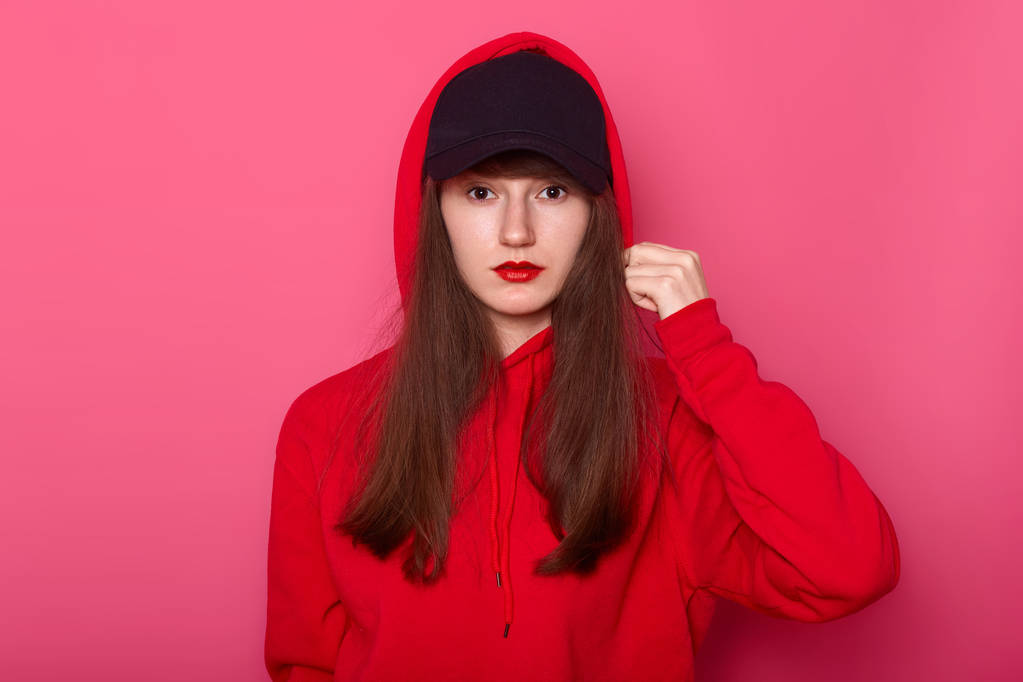 Image of brunette girl looking directly at camera with calm facial expression, wearing casual red hoody and black cap, touches her hood, posing isolated on rose background. Modeling concept. - Photo, Image