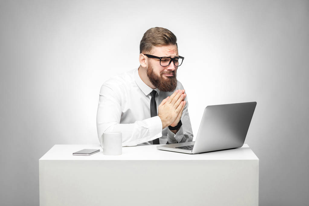Portrét nadějný vousatý mladý manažer v bílé košili a černé kravatě, modlil se a prohlížel si přenosný počítač při sezení na kancelářský stůl  - Fotografie, Obrázek