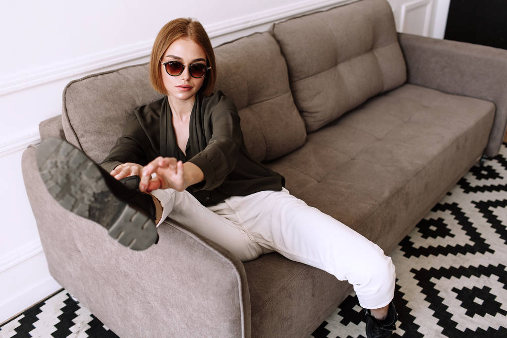 Beautiful sweet girl lying on the sofa. Golden stylish model with short red hair, plump lips, sunglasses, casual clothing, costume, outfit, overalls. Model test, advertising glasses, clothes - Photo, Image