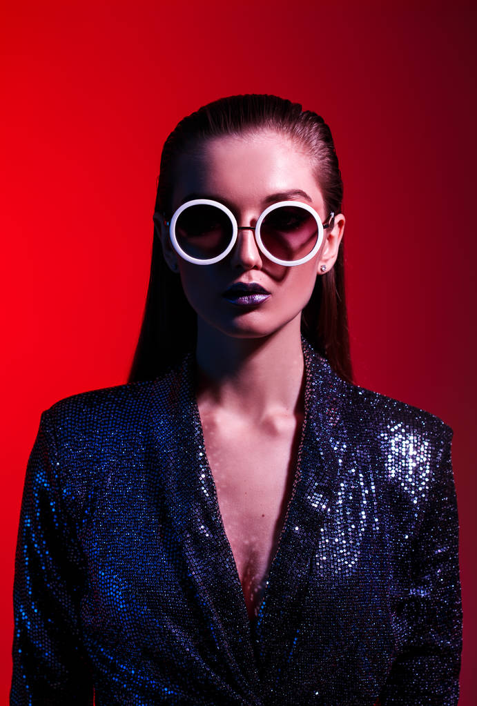 Moda dziewczyna z długimi włosami i okrągłe okulary przeciwsłoneczne w czarnym lśniącej sukience stanowi w neonowym świetle w studio - Zdjęcie, obraz