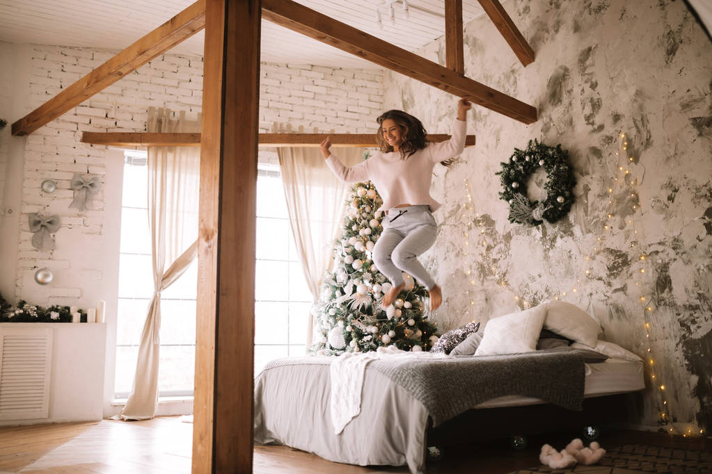 Viehättävä tyttö pukeutunut valkoinen villapaita ja housut hyppää sängyllä harmaa huopa ja valkoinen tyynyt viihtyisä sisustettu huone uudenvuoden puu
 - Valokuva, kuva