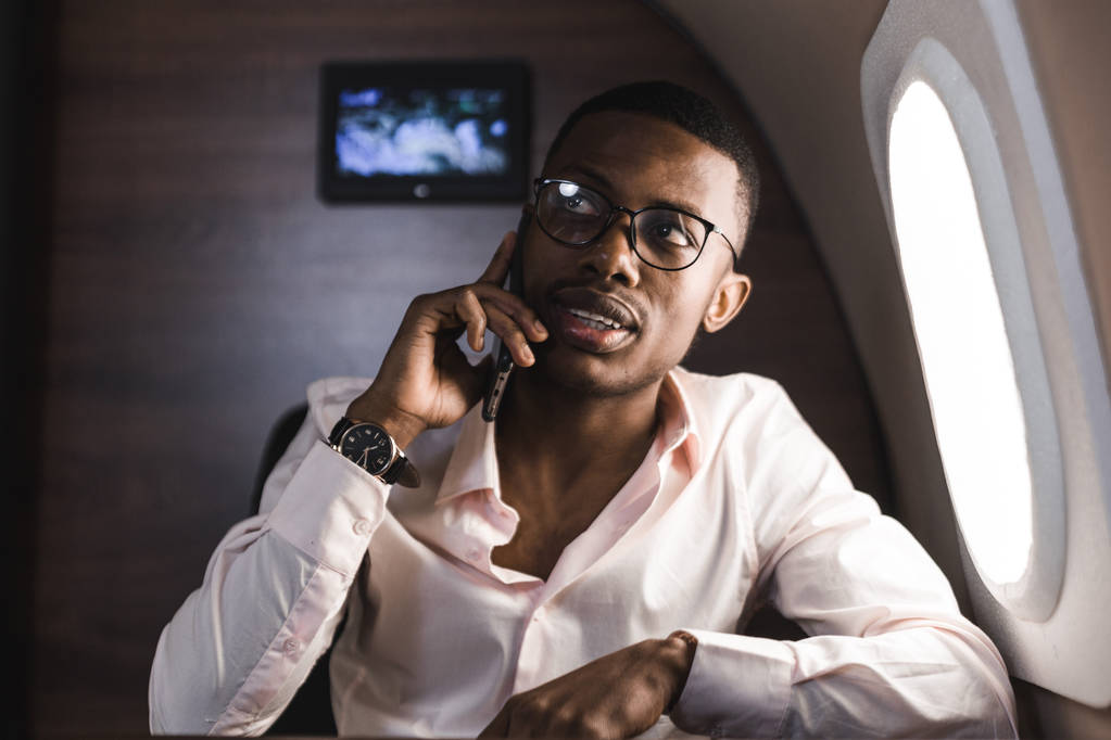 Giovane uomo d'affari afroamericano attraente e di successo con vetrate parlare al telefono e lavorare mentre seduto sulla sedia del suo aereo d'affari privato
 - Foto, immagini