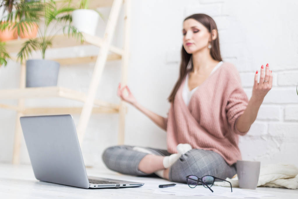 Joven mujer feliz se sienta en el suelo en una pose de yoga en un apartamento luminoso y trabaja detrás de un ordenador portátil, una chica freelancer medita mientras trabaja. Estrés del trabajo
 - Foto, imagen