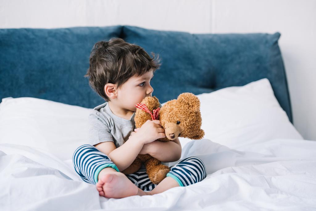 yalınayak yatakta oturan ve oyuncak ayı tutan sevimli çocuk  - Fotoğraf, Görsel