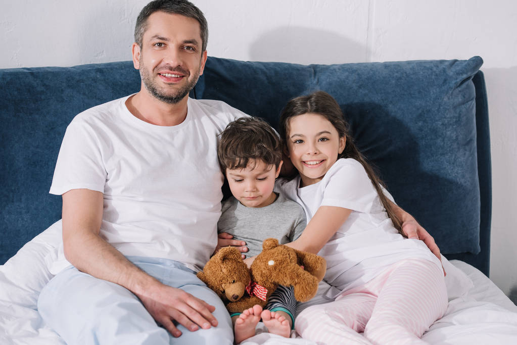 alegre padre sentado en la cama con feliz hija y triste hijo pequeño
  - Foto, imagen