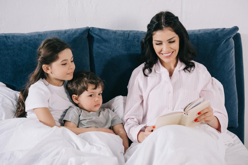 alegre madre leyendo libro cerca de linda hija e hijo en el dormitorio
  - Foto, Imagen