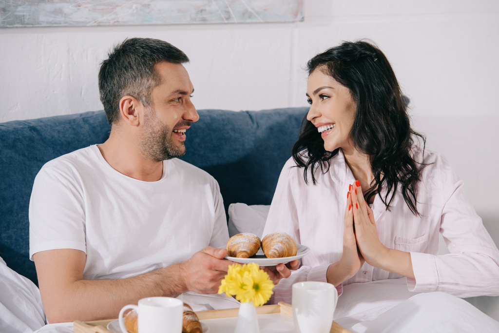 hombre alegre sosteniendo plato con croissants cerca de la mujer feliz en el dormitorio
  - Foto, Imagen