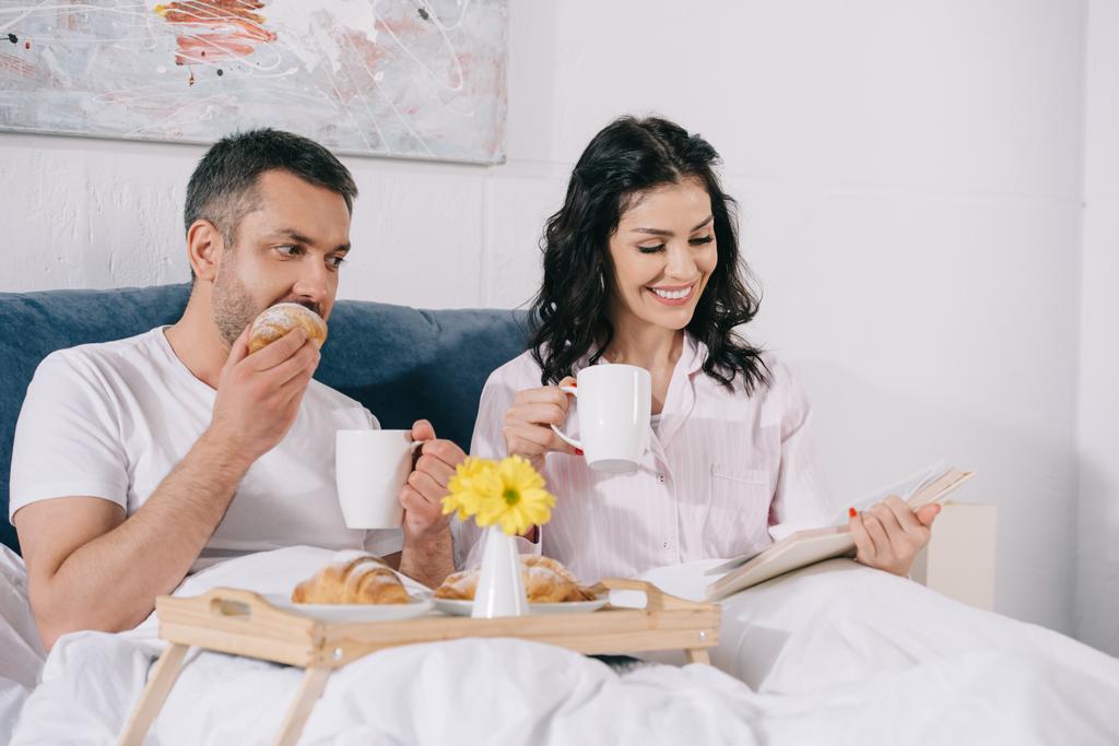 χαρούμενη γυναίκα διαβάζοντας βιβλίο κοντά στον σύζυγο τρώγοντας κρουασάν στο κρεβάτι  - Φωτογραφία, εικόνα