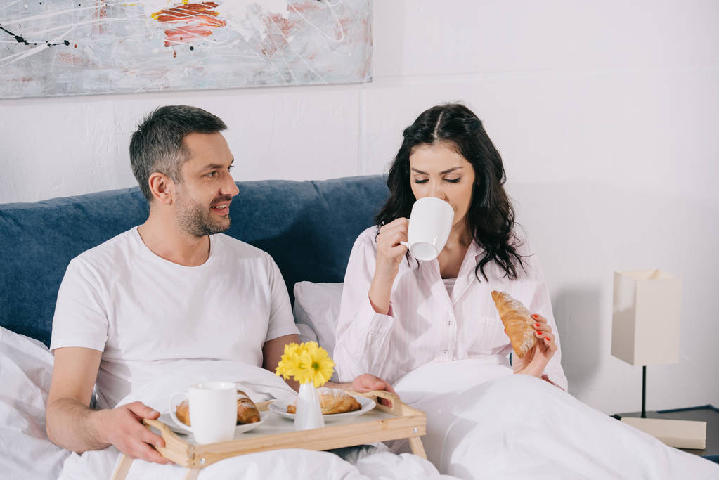 gelukkige man kijken naar aantrekkelijke vrouw drinken van koffie en het houden van croissant in bed  - Foto, afbeelding