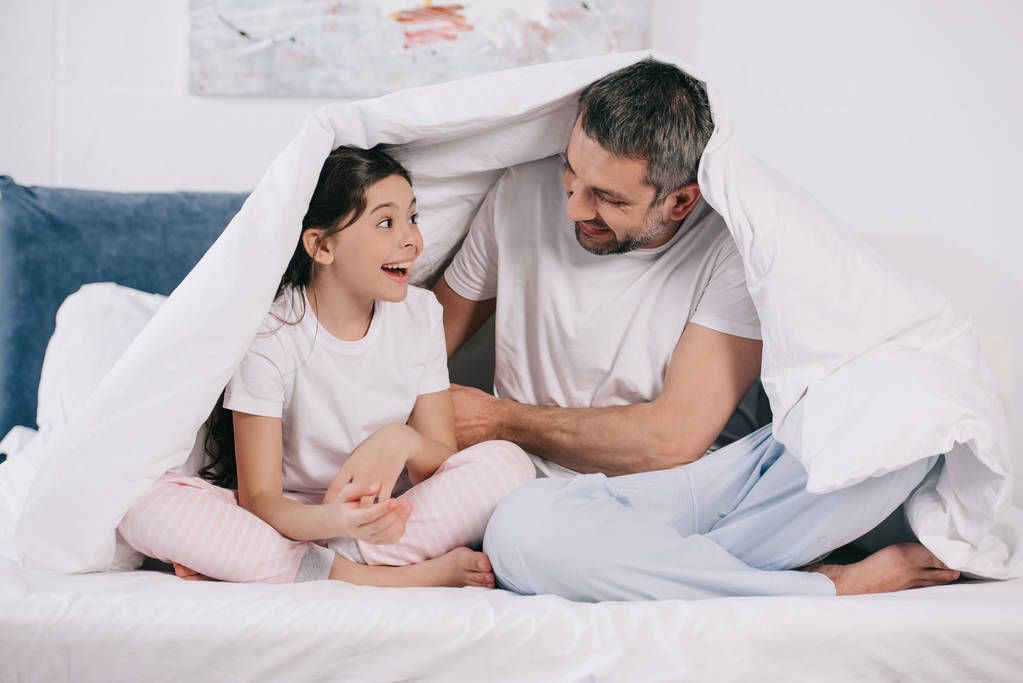 wesoły ojciec siedzi z szczęśliwą córką i trzyma koc na łóżku  - Zdjęcie, obraz