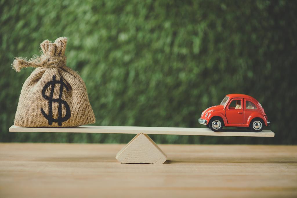 speelgoed auto en geld tas met dollarteken balanceren op wip op groene achtergrond - Foto, afbeelding