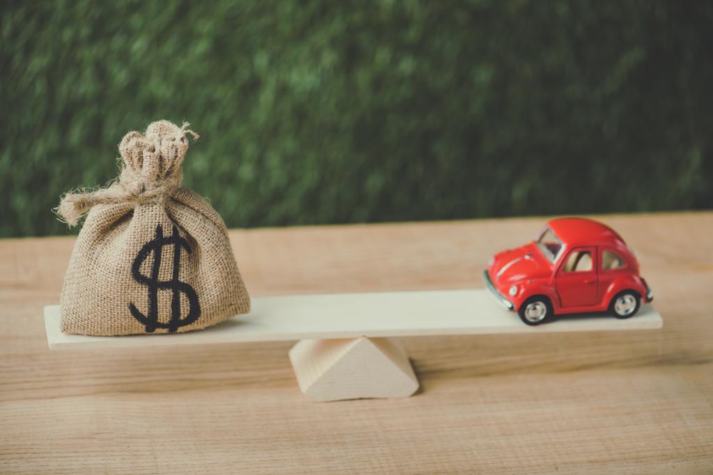 κόκκινο μινιατούρα αυτοκίνητο και τσάντα χρήματα με το σύμβολο του δολαρίου εξισορρόπηση στην τραμπάλα σε πράσινο φόντο - Φωτογραφία, εικόνα