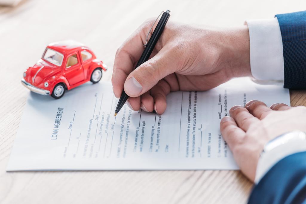 cropped view of car dealer writing in loan agreement near red toy car on wooden tabletop - Photo, Image