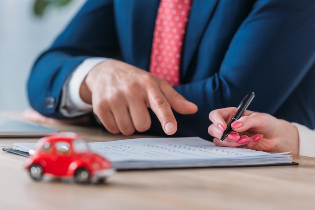gedeeltelijke weergave van de cliënt die de pen en de autodealer aanwijst op een handtekening plaats in een leningsovereenkomst in de buurt van rode speelgoed auto op tafel - Foto, afbeelding