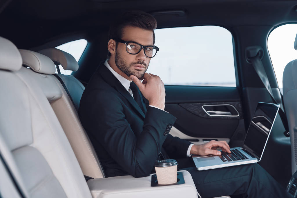 Successful professional. Thoughtful young man in full suit working using laptop and adjusting his eyewear while sitting in the car - Photo, Image