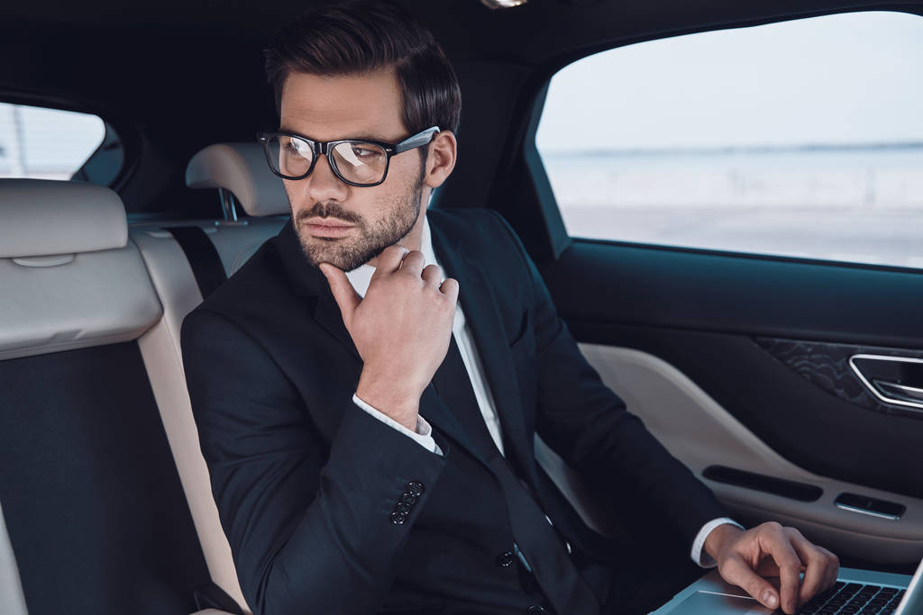 Myślenie o biznesie. Przemyślany młody człowiek w pełnym garnitur pracy przy użyciu laptopa i dostosowanie jego okulary siedząc w samochodzie   - Zdjęcie, obraz
