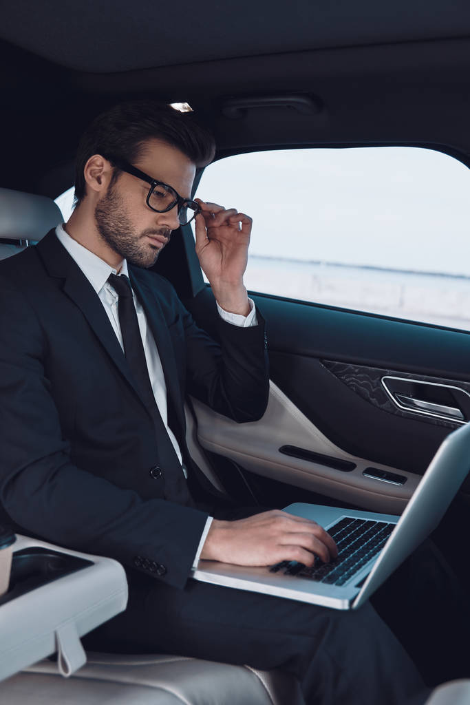 Byznys je jeho život. Přemýšlivý mladík v plném obleku pracující s laptopem, který si při sezení v autě nastavil brýle. - Fotografie, Obrázek