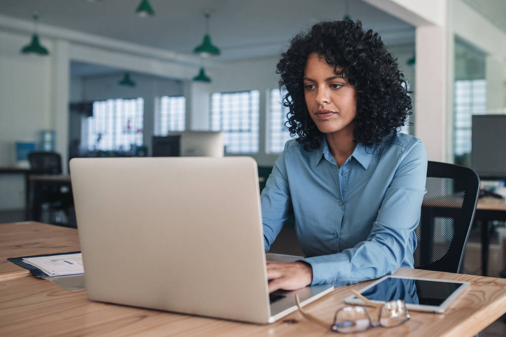 Kendini adamış olgun iş kadını bir dizüstü bilgisayar ile online çalışan büyük bir modern ofiste masasında oturan - Fotoğraf, Görsel