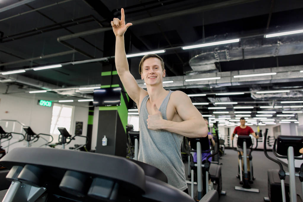 Junger Mann in Sportbekleidung läuft im Fitnessstudio auf Laufband und zeigt Daumen hoch - Foto, Bild