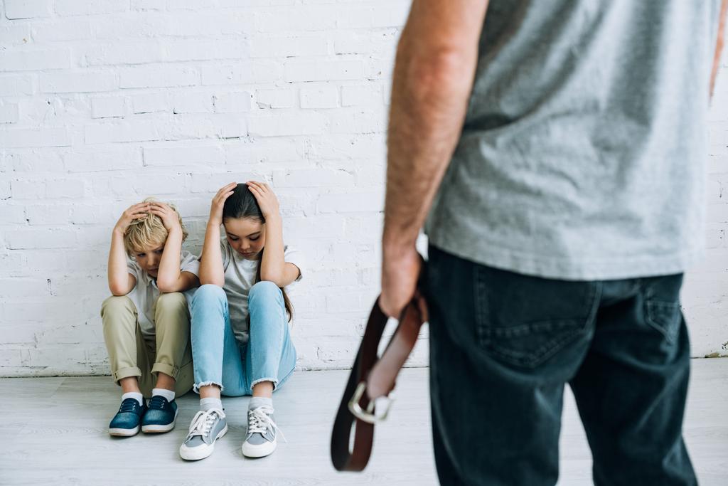 vista recortada de un padre abusivo sujetando el cinturón y niños asustados sentados en el suelo
 - Foto, imagen