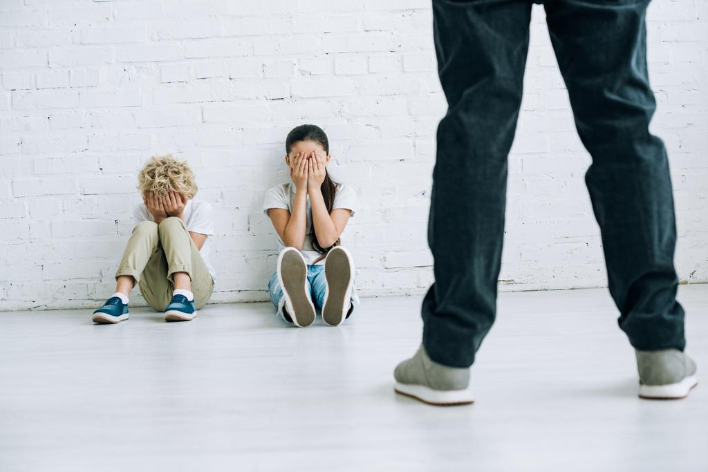 обрізаний вид на жорстокого батька, який тримає пояс і наляканих дітей, що сидять на підлозі
 - Фото, зображення