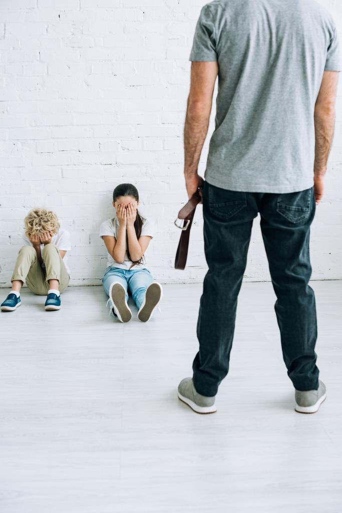 oříznutý pohled na urážlivého otce přidržující opasek a vystrašený děti sedící na podlaze - Fotografie, Obrázek