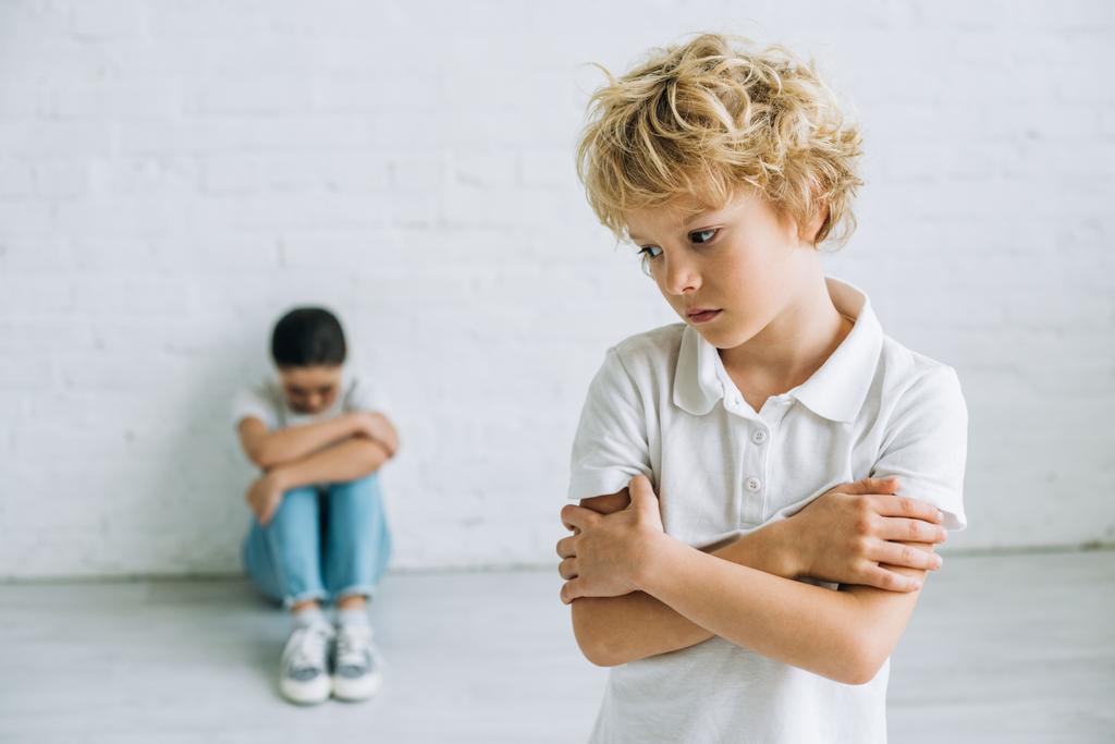 kız kardeşi yerde otururken ve evde ağlarken çapraz kolları ile duran üzgün çocuk - Fotoğraf, Görsel