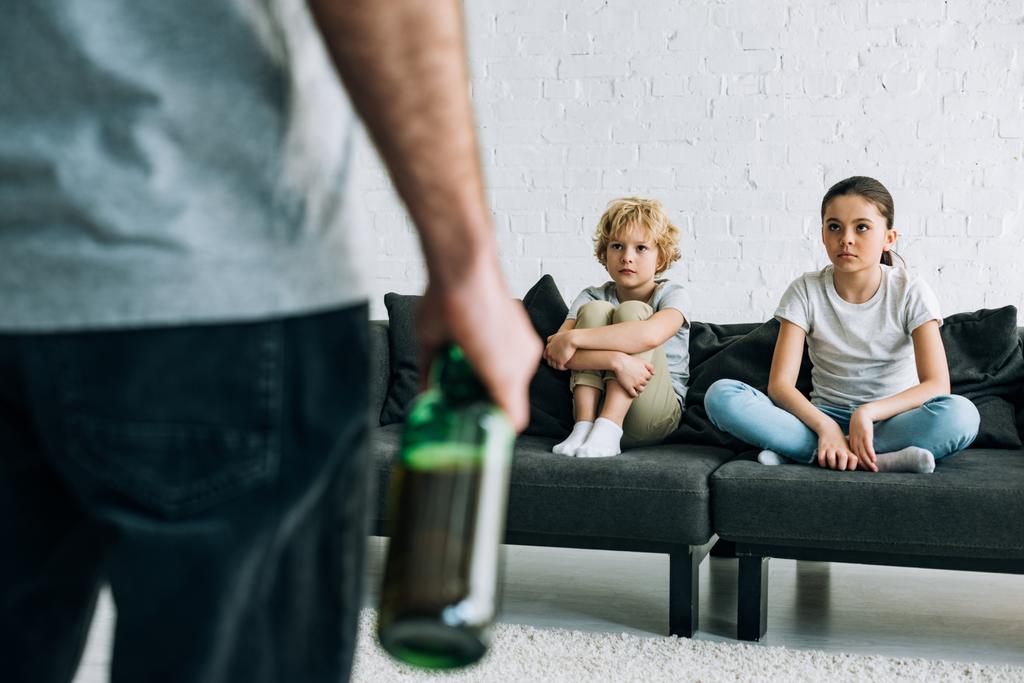 rajattu näkymä alkoholiriippuvaisesta isästä oluella ja järkyttyneillä lapsilla sohvalla
 - Valokuva, kuva
