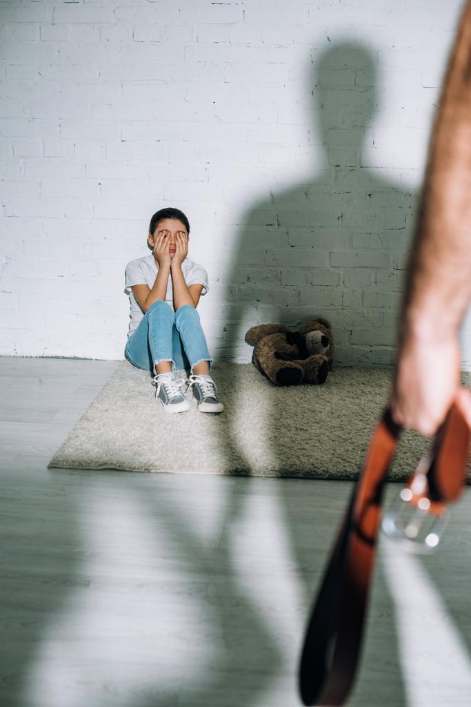 обрізаний вид батька тримає пояс і налякана дочка сидить на килимі біля його силуету
 - Фото, зображення