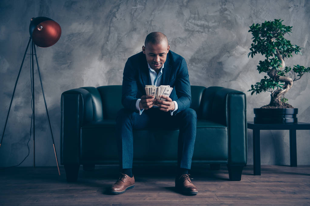 Портрет красивого стильного элегантного привлекательного занятого парня в костюме топ-менеджера, сидящего на диване, баланс бюджета на рабочем месте серая бетонная стена
 - Фото, изображение
