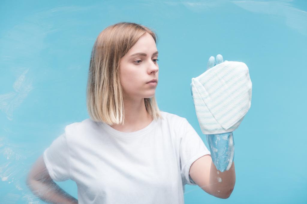 νεαρή γυναίκα σε λαστιχένιο γάντι καθαρισμού γυαλί με κουρέλι σε μπλε φόντο - Φωτογραφία, εικόνα