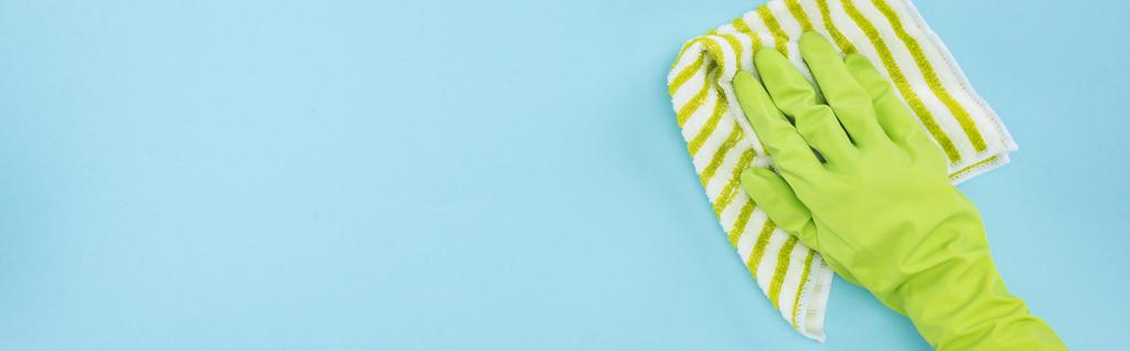 πανοραμική βολή καθαρότερο σε πράσινο ελαστικό γάντι κρατώντας ριγέ κουρέλι σε μπλε φόντο - Φωτογραφία, εικόνα