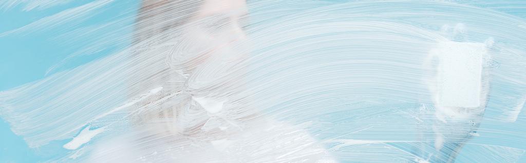 青い背景に白い泡とガラスの後ろにスポンジを持つ女性のパノラマショット - 写真・画像