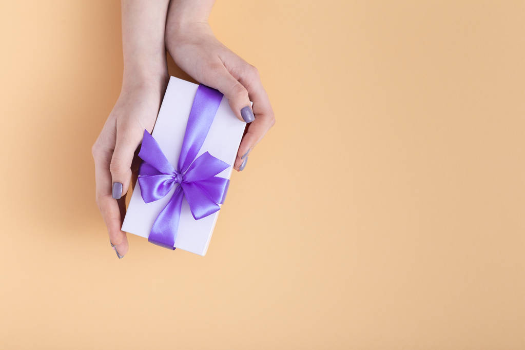 девушка, держащая в руках подарок, женщины с подарочной коробкой с привязанным лиловым бантом ленты в руках на пастельном оранжевом фоне, вид сверху, концептуальный праздник, любовь и забота
 - Фото, изображение