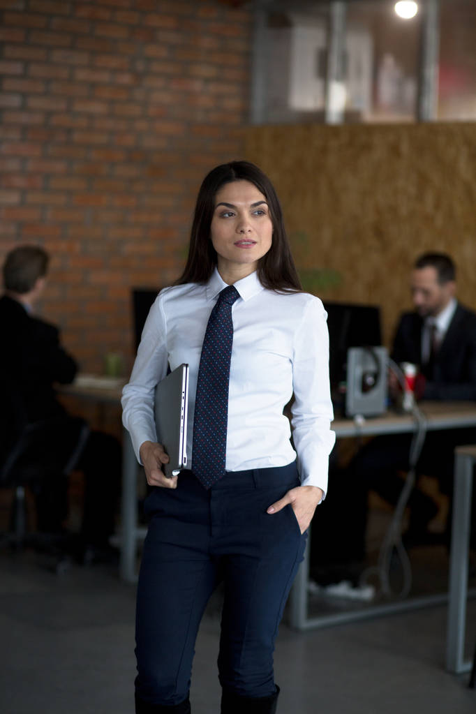 Pretty brunette is an office worker who is standing in office, holding a laptop, two colleguas are working behind her. - Photo, Image