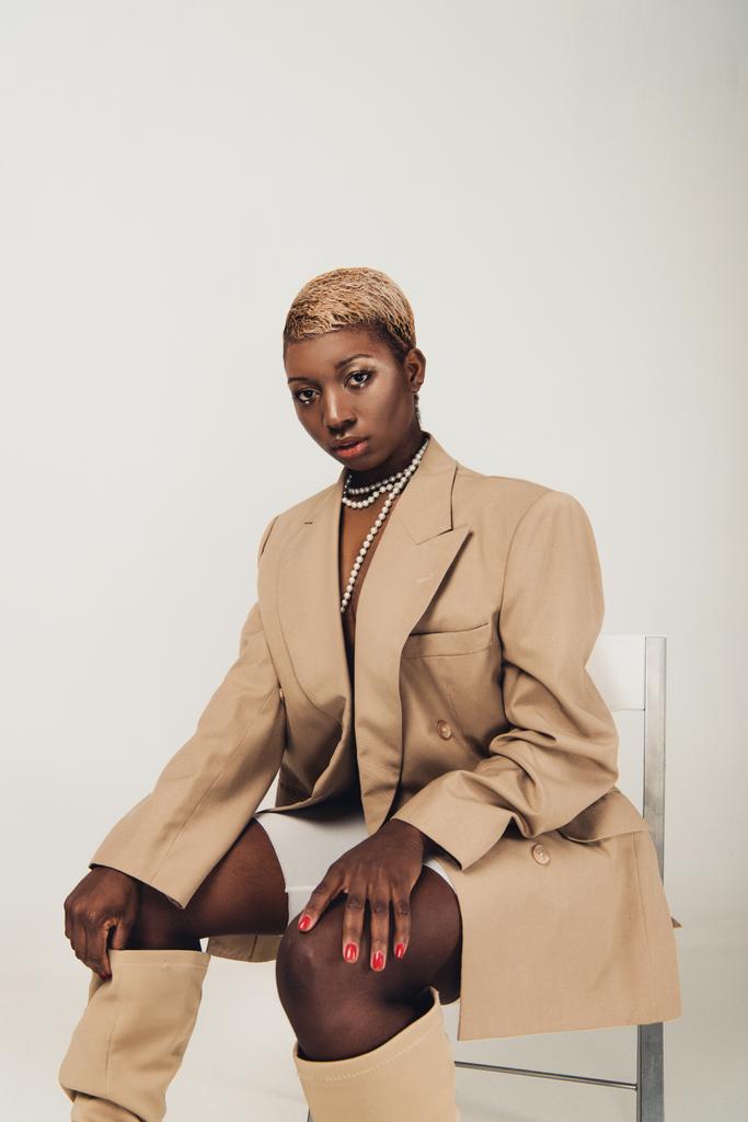 trendy african american woman in beige jacket and necklace sitting on chair isolated on grey - Photo, Image