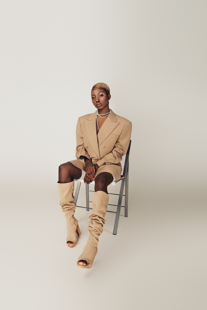 グレーの椅子に座っているベージュのジャケットのエレガントなアフリカ系アメリカ人モデル - 写真・画像