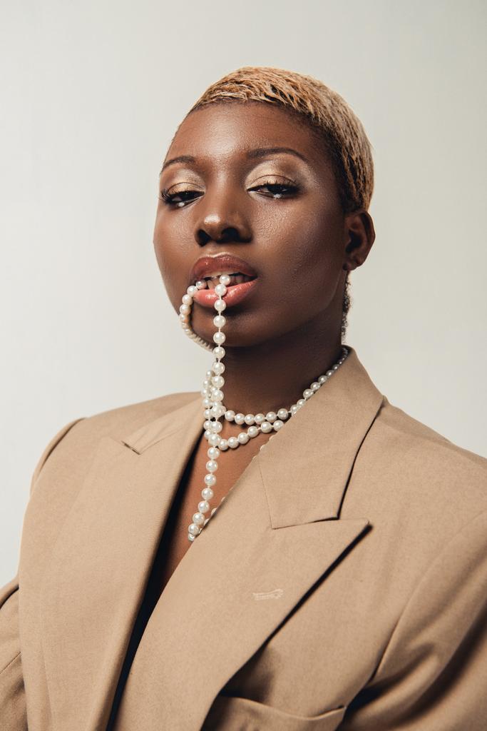 portrait of fashionable african american woman in beige jacket and necklace isolated on grey - Photo, Image