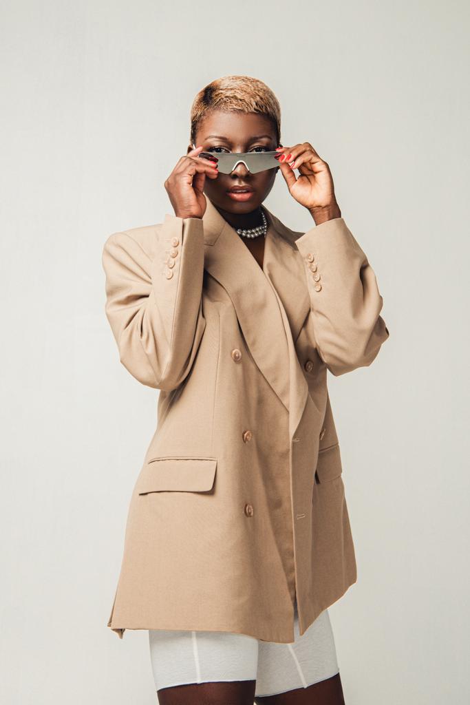tyylikäs afrikkalainen amerikkalainen tyttö poseeraa aurinkolasit ja tyylikäs beige takki eristetty harmaa
 - Valokuva, kuva