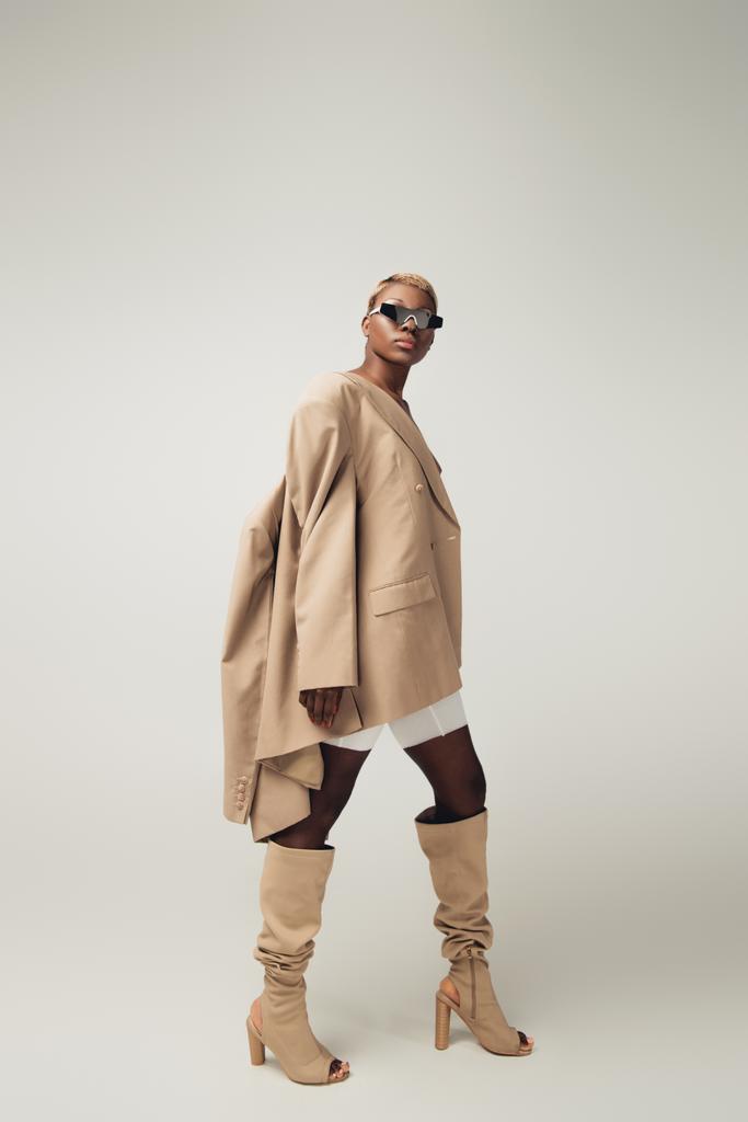 elegante joven afroamericana chica posando en gafas de sol y chaqueta beige en gris
 - Foto, Imagen