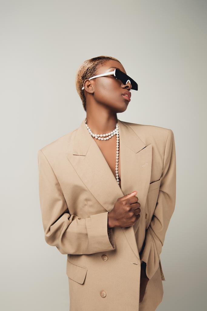 chica afroamericana elegante posando en gafas de sol y chaqueta beige elegante aislado en gris
 - Foto, imagen