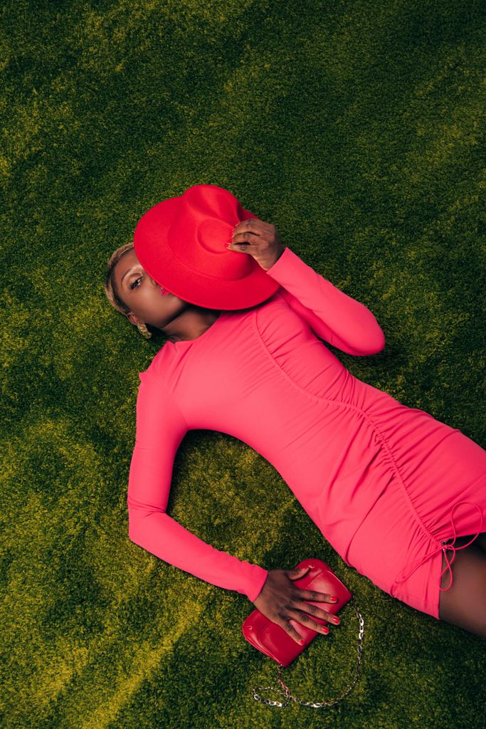 緑の草の上に帽子とバッグでポーズピンクのドレスで美しいアフリカ系アメリカ人女性のトップビュー - 写真・画像