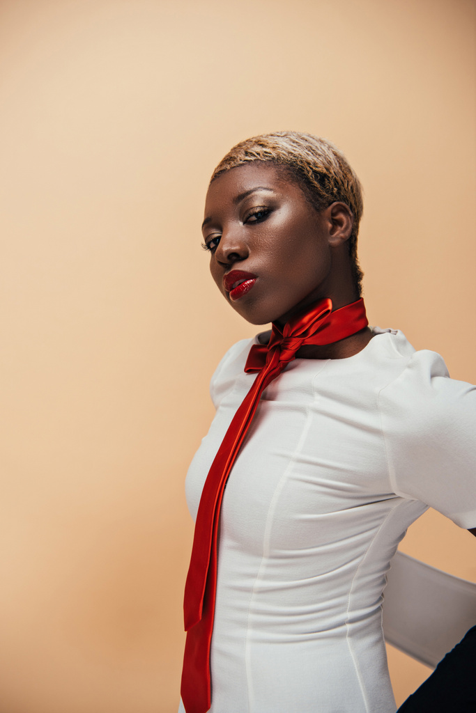 μοντέρνα αφρικανική αμερικανική μοντέλο που ποζάρει σε λευκό φόρεμα και κόκκινο κασκόλ απομονώνονται σε μπεζ - Φωτογραφία, εικόνα