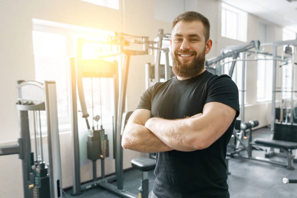 Portrét usmívajícího se osobního fitness trenéra v tělocvičně, sebevědomý muž se založenýma rukama na kameře. Fitness, sport, trénink, koncepce lidí - Fotografie, Obrázek
