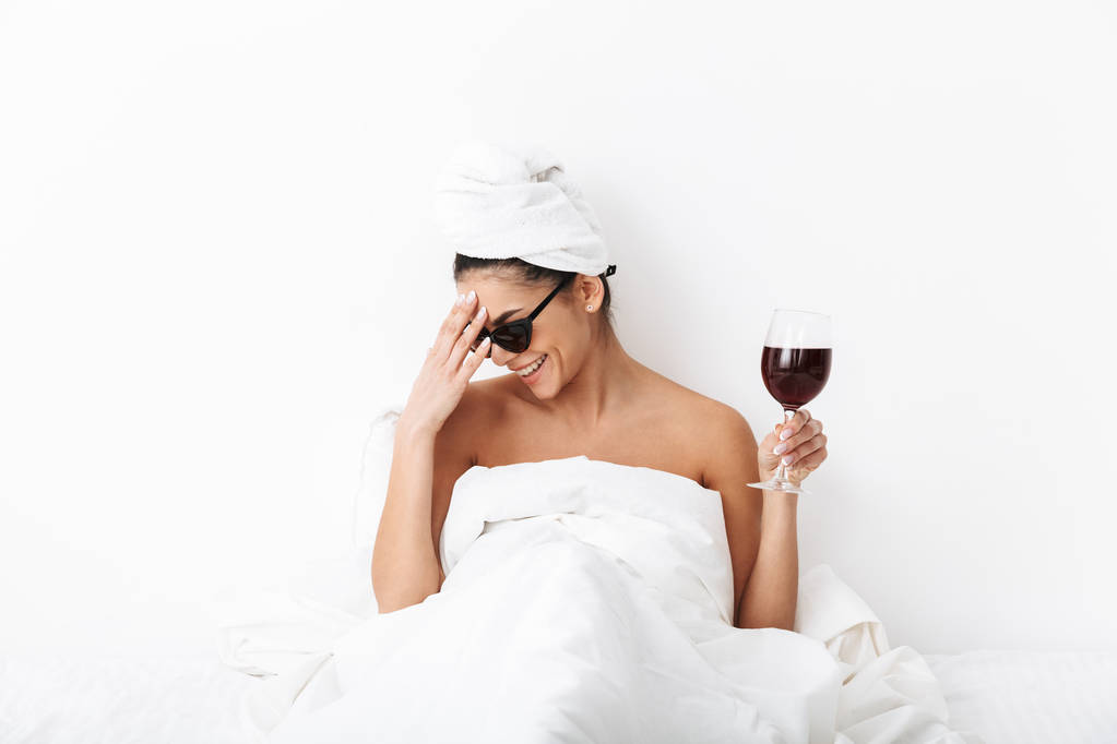 Lachen gelukkige vrouw met handdoek op hoofd ligt in bed onder deken geïsoleerd over witte muur achtergrond dragen zonnebril drinken wijn. - Foto, afbeelding