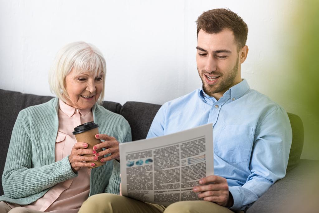 smiling senior mother holding coffee and reading newspaper with son at home - Photo, Image