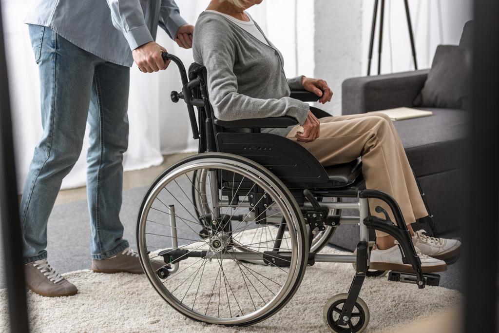 vista recortada del hombre que lleva a la madre discapacitada en silla de ruedas
 - Foto, Imagen