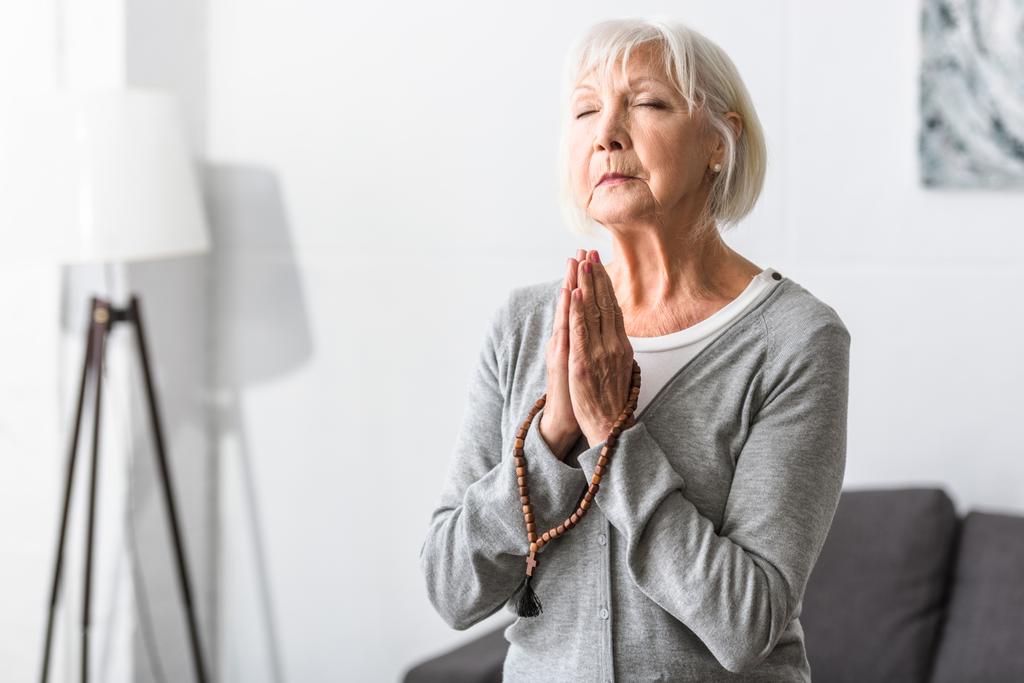 mujer mayor sosteniendo rosario de madera y orando con los ojos cerrados
 - Foto, imagen
