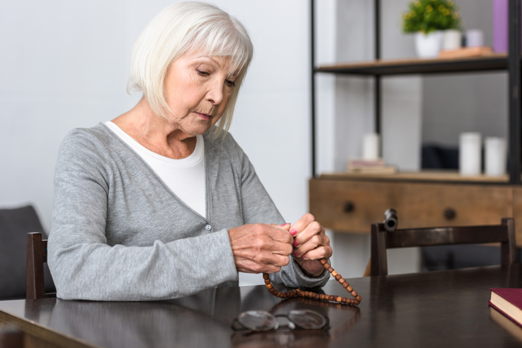 пенсійна старша жінка сидить за столом і тримає розарій
 - Фото, зображення