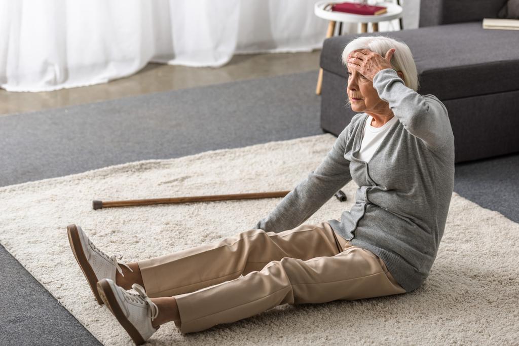 пожилая женщина с мигренью сидит на ковре и трогает лоб рукой
 - Фото, изображение